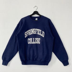 Puede incluir: Sudadera azul marino de Springfield College con letras blancas. El texto "SPRINGFIELD COLLEGE" está arqueado en el pecho. La sudadera está colgada de una percha negra sobre un fondo blanco.