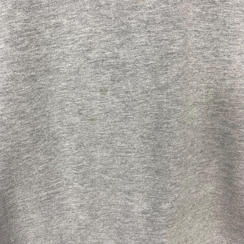 May include: Close-up of a heather gray fabric with a textured surface. The fabric has a few small, dark spots. The material appears to be a soft knit, possibly cotton or a cotton blend. The image is well-lit, showing the details of the textile.