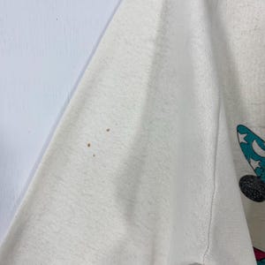 Pu&ograve; includere: Maglietta a maniche lunghe bianco sporco con grafica a cartoni animati. La grafica presenta un topo nero con un cappello blu decorato con stelle bianche e una luna crescente. La maglietta ha macchie marroni sulla manica.