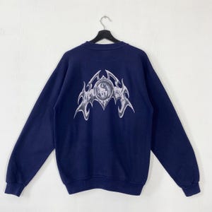 Puede incluir: Sudadera azul marino con un gran dise&ntilde;o gr&aacute;fico plateado estilizado en la espalda. El dise&ntilde;o presenta un emblema central rodeado de un dise&ntilde;o en forma de llama. La sudadera est&aacute; colgada en una percha negra sobre un fondo blanco.