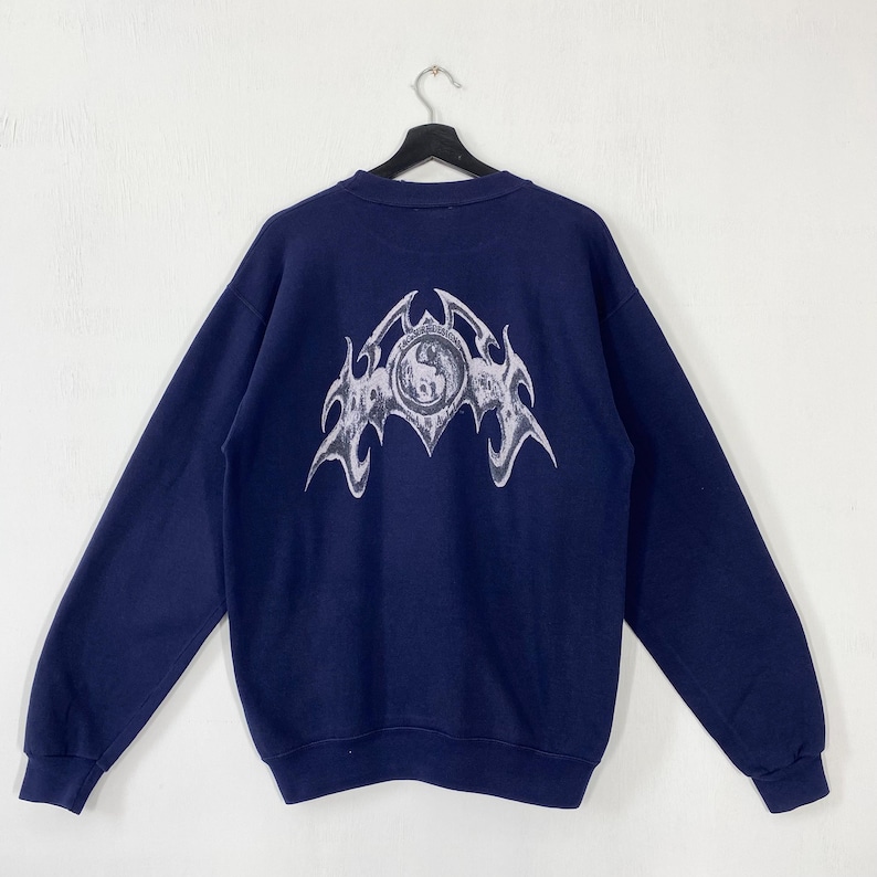 Puede incluir: Sudadera azul marino con un gran dise&ntilde;o gr&aacute;fico plateado estilizado en la espalda. El dise&ntilde;o presenta un emblema central rodeado de un dise&ntilde;o en forma de llama. La sudadera est&aacute; colgada en una percha negra sobre un fondo blanco.