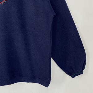 May include: Navy blue sweatshirt with embroidered floral detailing in red, blue, and green near the neckline. The long sleeves have a ribbed cuff with green and navy stripes. The sweatshirt is made of a soft, comfortable material.