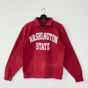 May include: A red sweatshirt featuring a quarter-zip collar and the white text "WASHINGTON STATE". The long-sleeved top has a relaxed fit. The brand name "Jansport" is visible on the collar. The sweatshirt is a classic style.