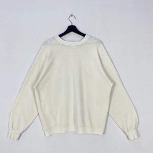 Pu&ograve; includere: Felpa a maniche lunghe color crema con scollo tondo, appesa a una gruccia nera. Le maniche hanno polsini a coste. Lo sfondo &egrave; bianco.