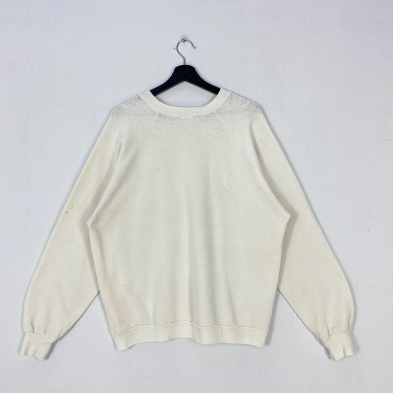 Pu&ograve; includere: Felpa a maniche lunghe color crema con scollo tondo, appesa a una gruccia nera. Le maniche hanno polsini a coste. Lo sfondo &egrave; bianco.