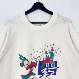 Pu&ograve; includere: Felpa color crema con una grafica di Topolino in abito da mago, che indica un castello. Il design include il testo "35 YEARS OF MAGIC" in uno stile colorato e vintage. La felpa ha un girocollo e maniche lunghe.