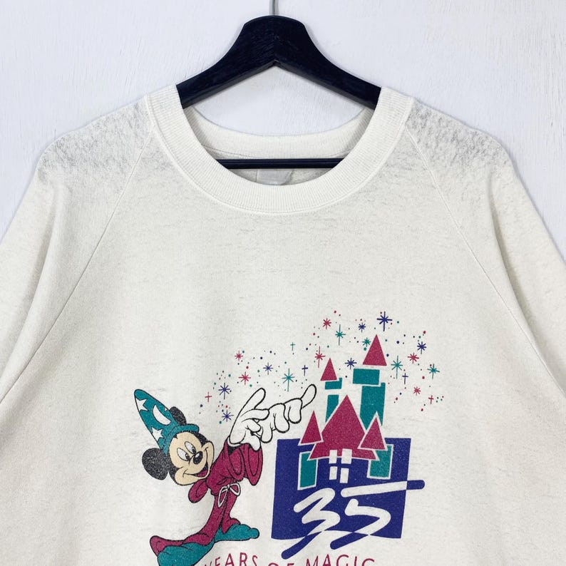 Pu&ograve; includere: Felpa color crema con una grafica di Topolino in abito da mago, che indica un castello. Il design include il testo "35 YEARS OF MAGIC" in uno stile colorato e vintage. La felpa ha un girocollo e maniche lunghe.