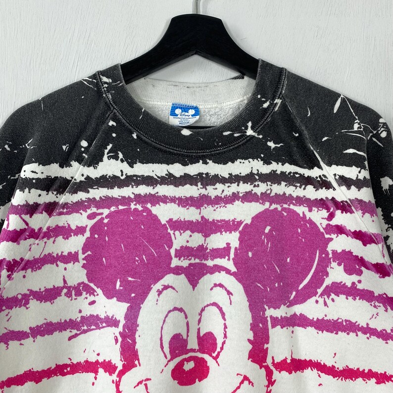 May include: White, pink, and black Mickey Mouse sweatshirt. The sweatshirt features a large Mickey Mouse graphic in pink, with a striped pattern in pink and white. The top of the sweatshirt is black with white paint splatters.
