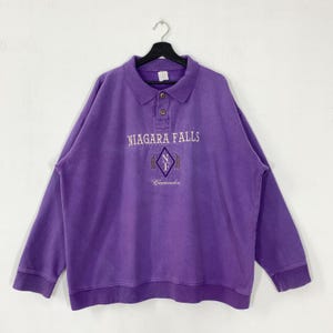 May include: A purple long-sleeved sweatshirt with a collared neckline and three buttons. The front features embroidered text that reads "NIAGARA FALLS" above a diamond-shaped emblem and the word "Canada".