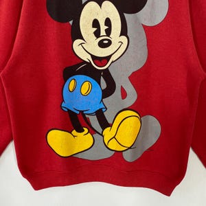 May include: Red sweatshirt with a large Mickey Mouse graphic. The cartoon character is in black, white, yellow, and blue. A grey outline of Mickey's head is visible behind the character.