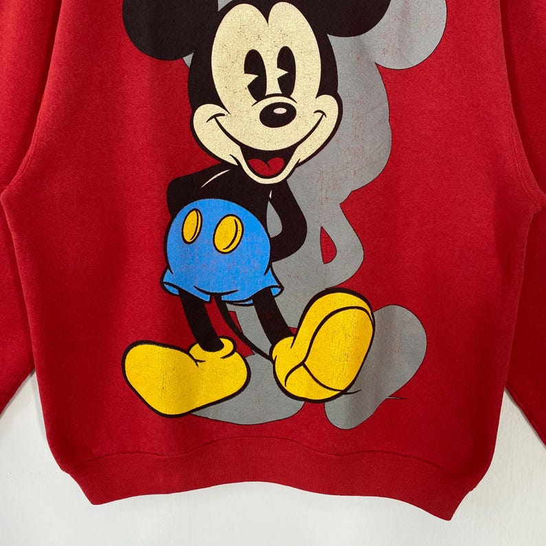 May include: Red sweatshirt with a large Mickey Mouse graphic. The cartoon character is in black, white, yellow, and blue. A grey outline of Mickey's head is visible behind the character.