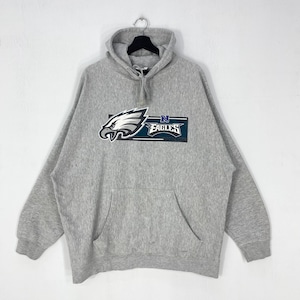 Può includere: Una felpa con cappuccio grigia con tasca frontale e coulisse. La felpa presenta una grafica di un'aquila bianca e verde acqua con la scritta "Eagles" in un design rettangolare. La felpa con cappuccio è appesa a una gruccia.
