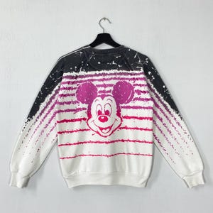 May include: White sweatshirt featuring a large Mickey Mouse graphic in shades of pink. The design includes horizontal pink stripes and a black and white gradient on the shoulders. A vintage Disney-themed apparel item.