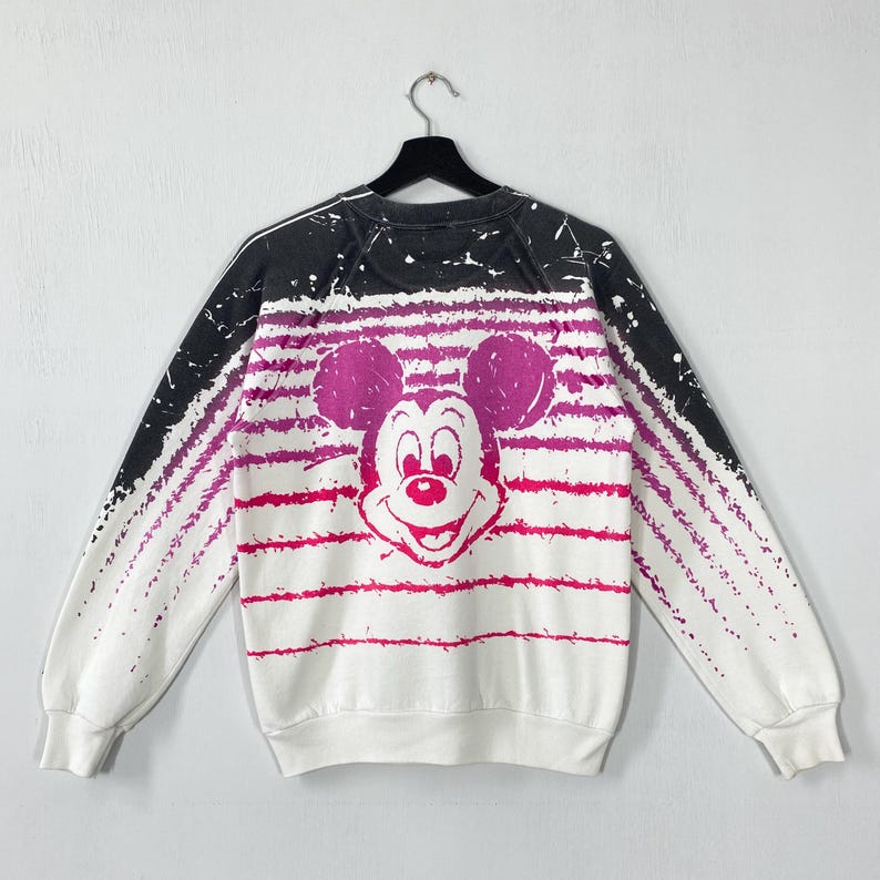 May include: White sweatshirt featuring a large Mickey Mouse graphic in shades of pink. The design includes horizontal pink stripes and a black and white gradient on the shoulders. A vintage Disney-themed apparel item.