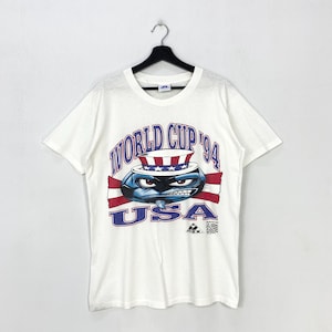 May include: White t-shirt featuring a cartoon character wearing a patriotic hat and sunglasses, with the text "WORLD CUP '94 USA" in blue and red. The shirt has a crew neck and short sleeves.