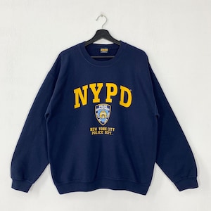 Peut inclure: Sweat-shirt bleu marine NYPD avec lettrage jaune et un emblème de la police. Le sweat-shirt a des manches longues et un col rond. Le texte "NEW YORK CITY POLICE DEPT." est en dessous de l'emblème.