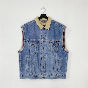 Può includere: Un gilet di jeans blu senza maniche con colletto in velluto a coste beige e fodera a quadri rossi e neri. Il gilet ha chiusure con bottoni, due tasche sul petto e due tasche laterali. Il logo Levi's è visibile.