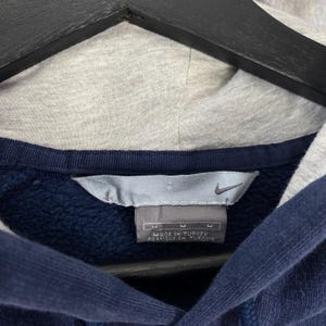 May include: Close-up of a navy blue hoodie with a gray hood. The Nike logo is visible on a tag that reads "Made in Turkey". The hoodie has a medium size.