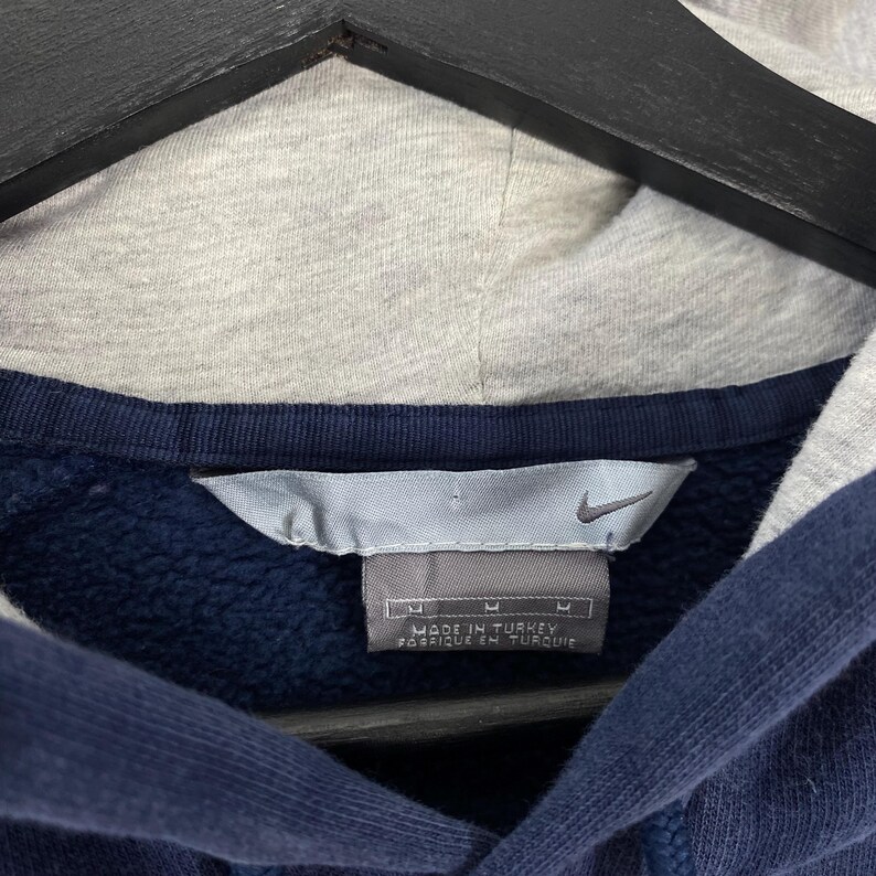 May include: Close-up of a navy blue hoodie with a gray hood. The Nike logo is visible on a tag that reads "Made in Turkey". The hoodie has a medium size.