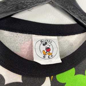 May include: Close-up of a vintage sweatshirt with a black collar and a tag featuring Mickey Mouse. The sweatshirt has a pattern of black, green, red, and yellow Mickey Mouse shapes. The tag reads "Mickey & Co."