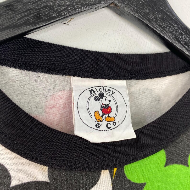 May include: Close-up of a vintage sweatshirt with a black collar and a tag featuring Mickey Mouse. The sweatshirt has a pattern of black, green, red, and yellow Mickey Mouse shapes. The tag reads "Mickey & Co."