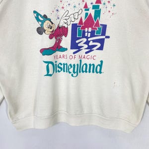 Pu&ograve; includere: Felpa color crema con una grafica di Topolino in abito da mago, con la scritta "Disneyland" e "35 Years of Magic". Il design include un castello e dettagli colorati. La felpa ha girocollo e maniche lunghe.