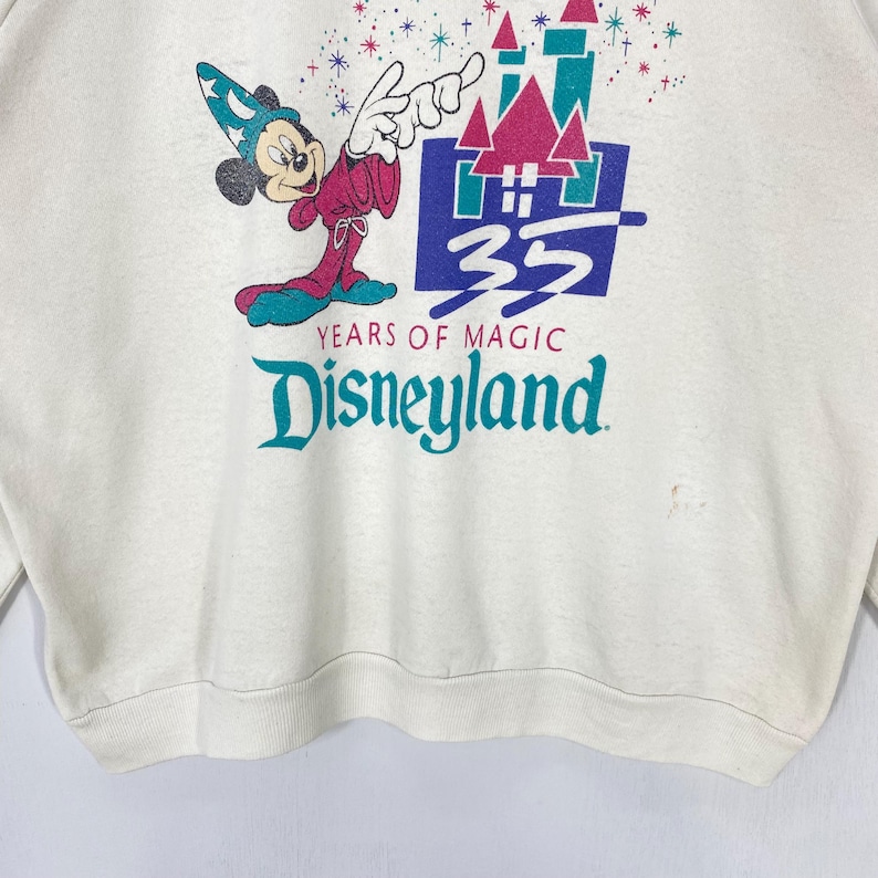 Pu&ograve; includere: Felpa color crema con una grafica di Topolino in abito da mago, con la scritta "Disneyland" e "35 Years of Magic". Il design include un castello e dettagli colorati. La felpa ha girocollo e maniche lunghe.