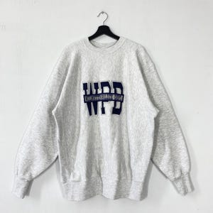 May include: A light grey sweatshirt with long sleeves, displaying the text "WPD WILMETTE POLICE DEPT" in navy blue. The sweatshirt is suspended from a black hanger against a white backdrop. The fabric appears to be soft and comfortable.