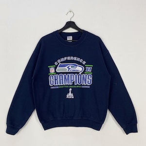 Pode incluir: Camisola azul marinho com a inscrição "CONFERENCE CHAMPIONS" e o logótipo dos Seattle Seahawks. O gráfico inclui o logótipo da NFL e o algarismo romano XLVIII. A camisola tem mangas compridas e gola redonda.