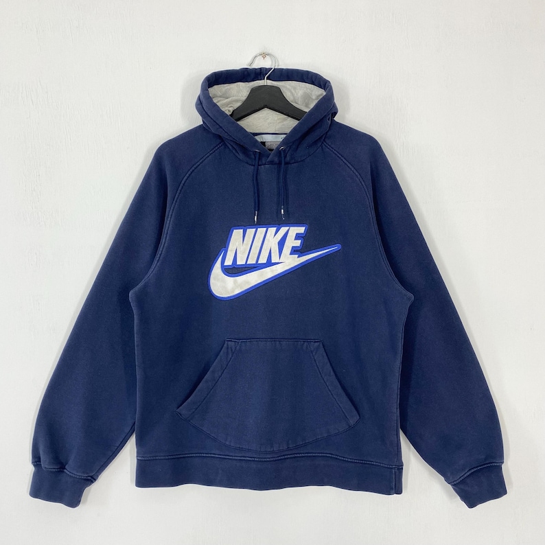 May include: Navy blue hoodie with a white and silver Nike logo on the front. The hoodie has a drawstring hood, a kangaroo pocket, and long sleeves. The interior of the hood is light gray.