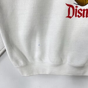 Puede incluir: Sudadera blanca con la palabra "Disney" en escritura roja. La sudadera tiene cuello redondo y mangas largas. La tela parece ser un material suave y c&oacute;modo.