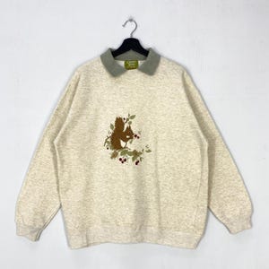 May include: A heathered beige sweatshirt featuring a sage green collar and a central embroidered graphic of a brown squirrel perched on a branch with green leaves and small red berries. The garment has long sleeves and a ribbed hem and cuffs.