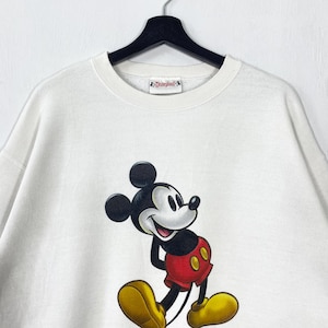 Puede incluir: Sudadera blanca de cuello redondo con un gran gr&aacute;fico de Mickey Mouse. Mickey lleva pantalones cortos rojos, zapatos amarillos y guantes negros. La sudadera tiene una etiqueta de Disneyland.