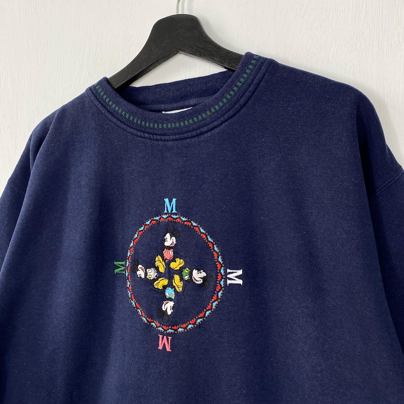 May include: Navy blue sweatshirt with a crew neck and a colorful embroidered design. The design features a circular pattern with the letter "M" at each cardinal point and a Mickey Mouse-inspired motif in the center.