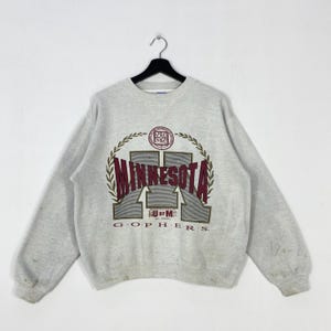 May include: A light gray sweatshirt with a crew neck, featuring a large graphic of "MINNESOTA" in maroon and gray, with "U OF M" and "GOPHERS" below. The design is framed by a laurel wreath. The sweatshirt is hanging on a black hanger.