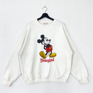 Puede incluir: Sudadera blanca con un gr&aacute;fico de Mickey Mouse en rojo, amarillo y negro. La palabra "Disneyland" est&aacute; impresa en rojo debajo del personaje. La sudadera est&aacute; colgada de una percha negra. Un dise&ntilde;o cl&aacute;sico de Disney.