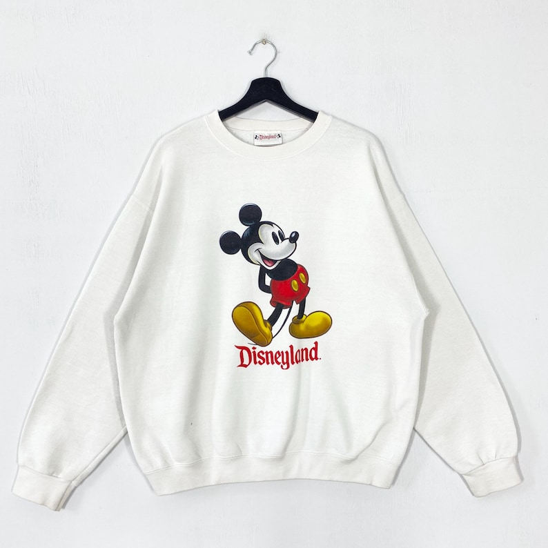 Puede incluir: Sudadera blanca con un gr&aacute;fico de Mickey Mouse en rojo, amarillo y negro. La palabra "Disneyland" est&aacute; impresa en rojo debajo del personaje. La sudadera est&aacute; colgada de una percha negra. Un dise&ntilde;o cl&aacute;sico de Disney.