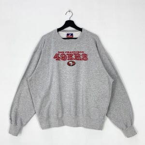 May include: A heather grey crewneck sweatshirt featuring the text SAN FRANCISCO 49ERS in red embroidered lettering across the chest, with a small 49ers team logo emblem positioned directly below the text and a Majestic brand label at the neckline.