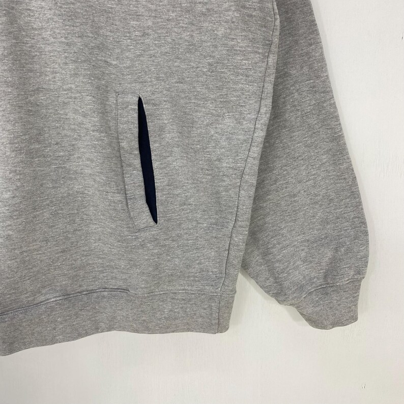 May include: A close-up of a gray sweatshirt with a dark blue pocket. The sweatshirt has a ribbed cuff and a simple, classic design. The fabric appears to be a soft, comfortable material.