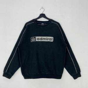 May include: A black long-sleeve sweatshirt featuring a crew neckline and ribbed cuffs. The chest displays a rectangular patch with the text admiral and a small logo. Thin white piping runs along the outer length of each sleeve from the shoulder.