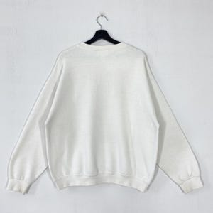 Puede incluir: Una sudadera blanca de cuello redondo con mangas largas, colgada de una percha negra sobre un fondo blanco. La sudadera tiene un dise&ntilde;o simple y cl&aacute;sico, adecuada para uso casual.