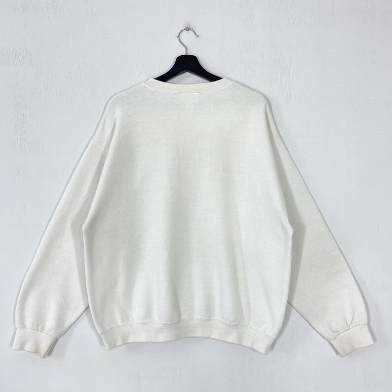 Puede incluir: Una sudadera blanca de cuello redondo con mangas largas, colgada de una percha negra sobre un fondo blanco. La sudadera tiene un dise&ntilde;o simple y cl&aacute;sico, adecuada para uso casual.