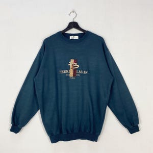 May include: Teal-colored sweatshirt with long sleeves, hanging on a hanger. The front features an embroidered logo with the text "PIERRE BALMAIN COLLECTION PARIS". The logo is in shades of brown, red, and gold.