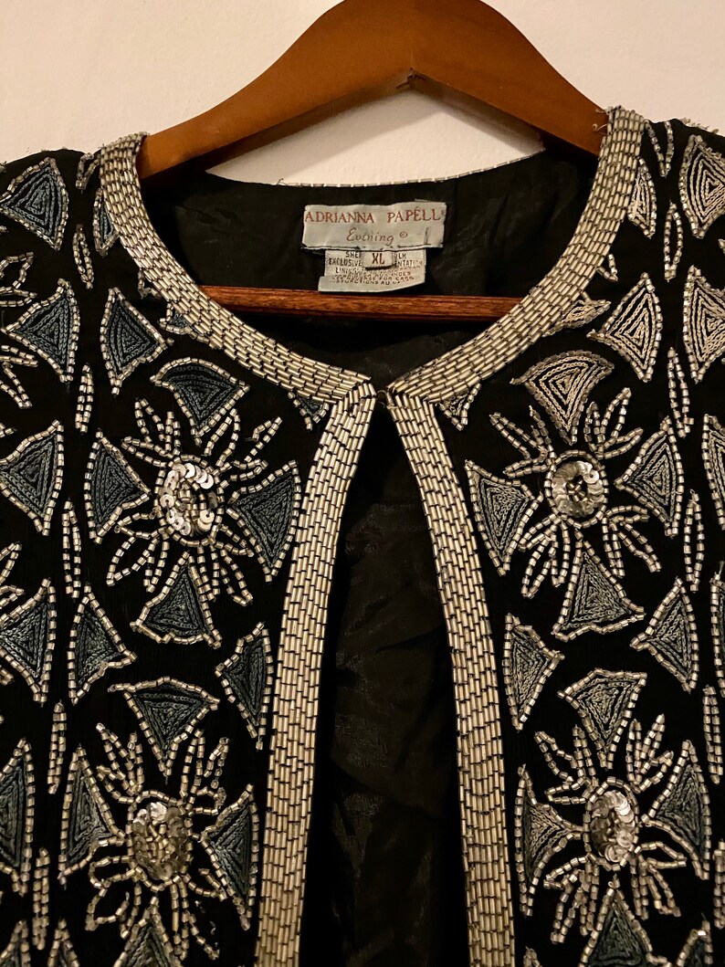 May include: A black beaded jacket with silver and blue beading in a geometric pattern. The label reads "Adrianna Papell Evening" and "Size XL".