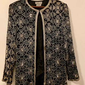 May include: A black beaded jacket with a silver and blue geometric pattern. The jacket has a front closure and long sleeves.