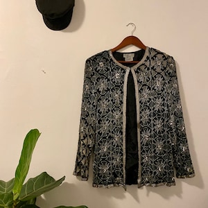 May include: A black sequined jacket with a silver floral pattern. The jacket has a long sleeve and a open front.