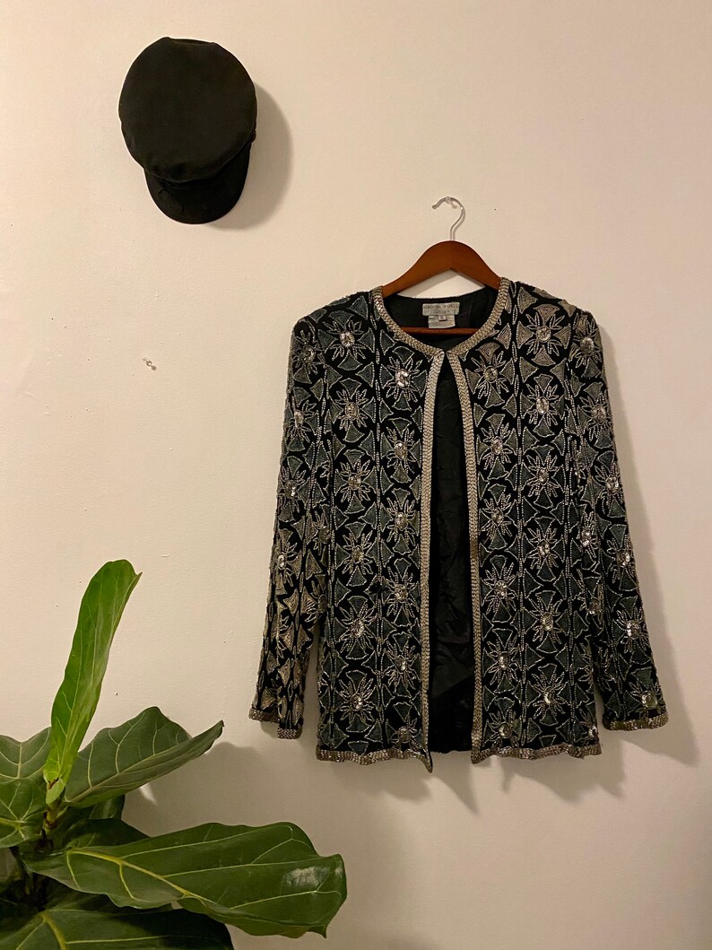 May include: A black sequined jacket with a silver floral pattern. The jacket has a long sleeve and a open front.