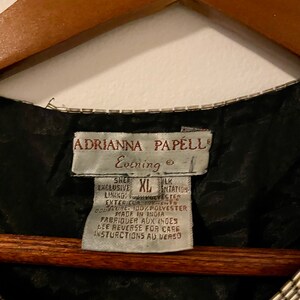 May include: Black evening dress with a silver beaded neckline and a white tag that reads "Adrianna Papell Evening" and "Sher Exclusive XL Silk Intation Lining 100% Polyester Exterieur 100% Polyester Doublure Made in India Fabriquer aux Indes See Reverse for Care Instructions au Verso"