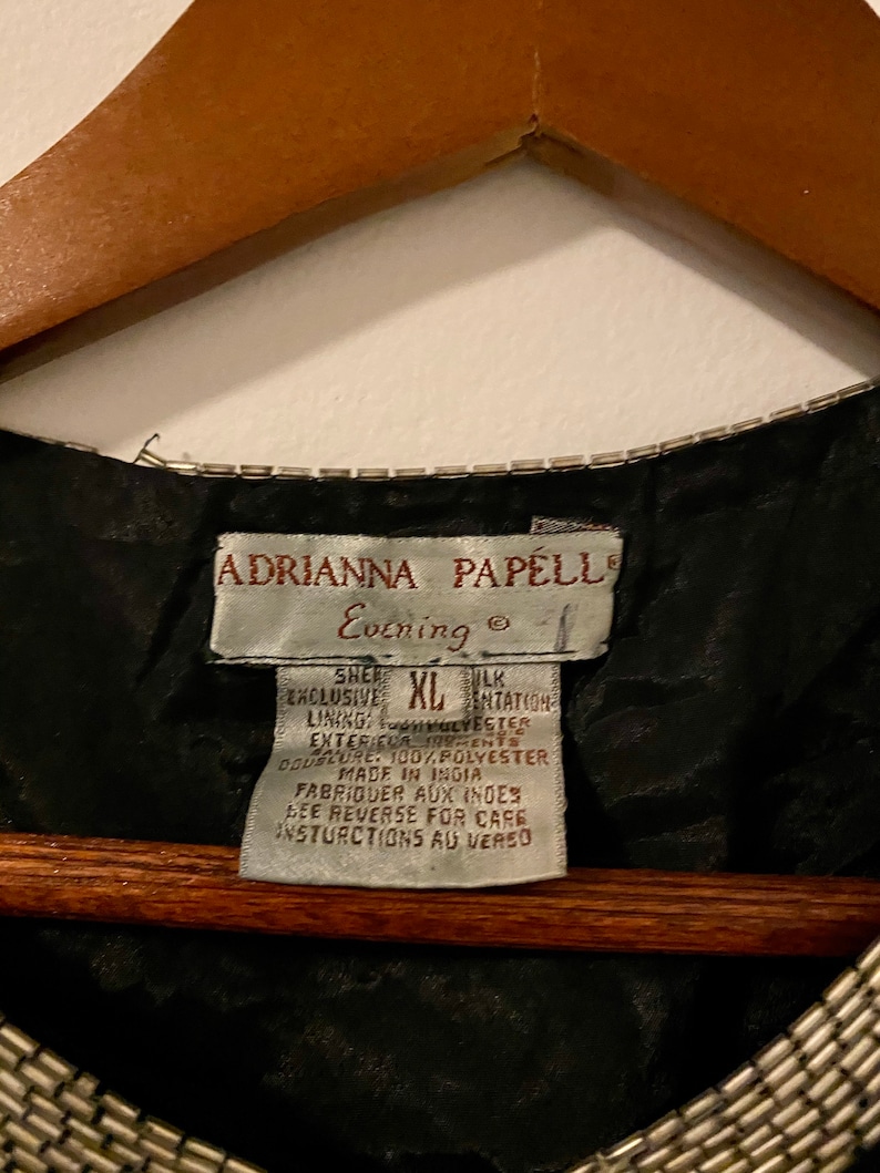 May include: Black evening dress with a silver beaded neckline and a white tag that reads "Adrianna Papell Evening" and "Sher Exclusive XL Silk Intation Lining 100% Polyester Exterieur 100% Polyester Doublure Made in India Fabriquer aux Indes See Reverse for Care Instructions au Verso"