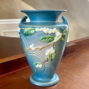 May include: A blue ceramic vase featuring a raised relief design of brown branches with green leaves and white blossoms. The vase has a flared rim, two small side handles, and a tapered body with subtle curved line detailing on the surface.